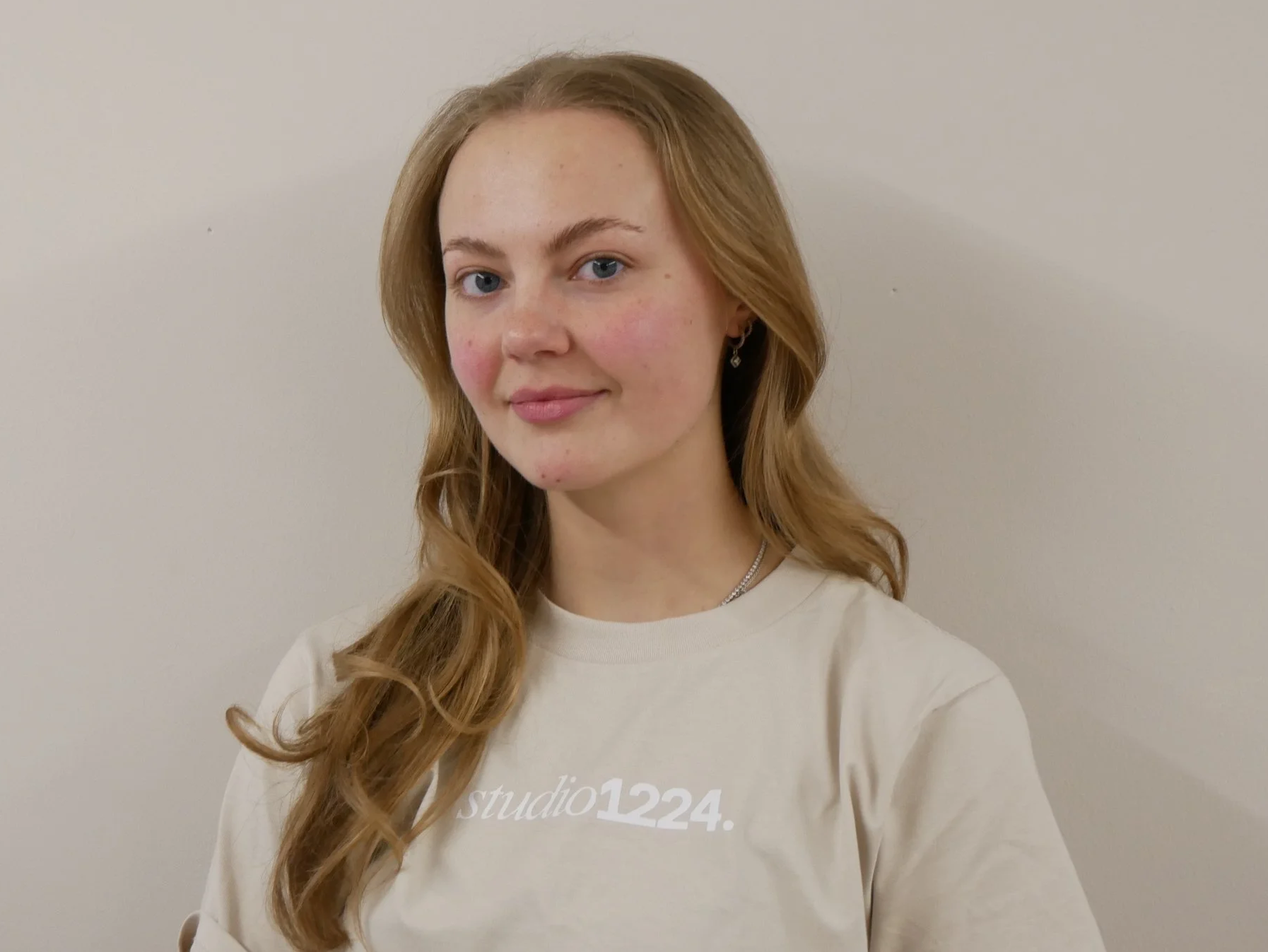 A young woman with long, wavy auburn hair, blue eyes, and light skin, smiling subtly against a plain off-white wall. She is wearing earrings, a silver necklace, and a beige sweatshirt with partially visible white text.