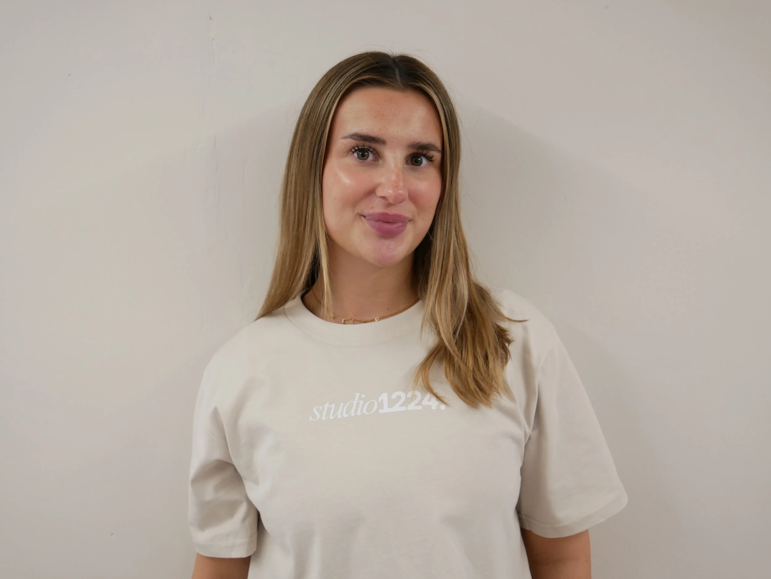 A young woman with long, light brown hair and light makeup, wearing a white sweatshirt with text on it, standing against a plain light-colored wall.