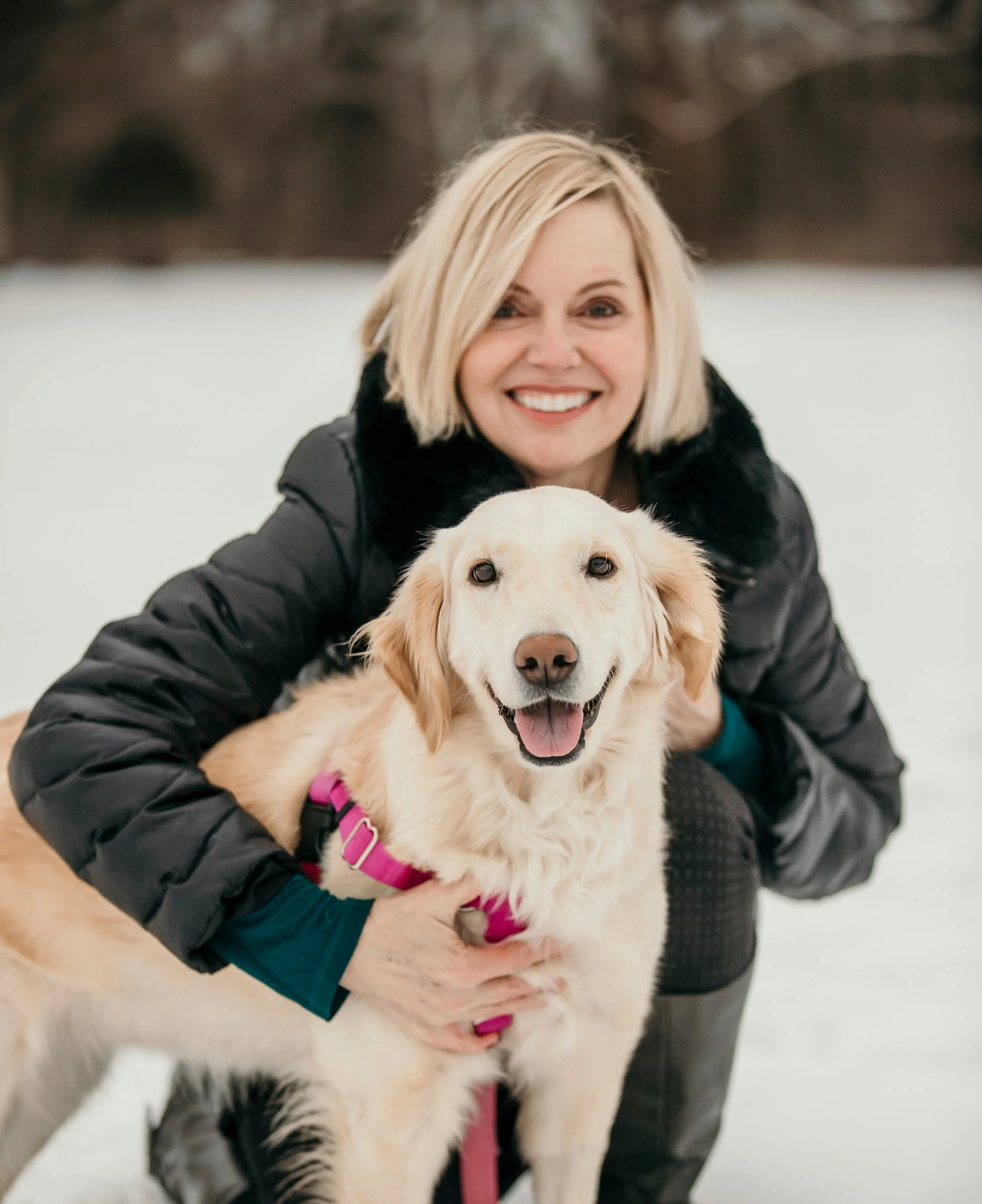 A woman with blonde hair wearing a black jacket and teal long sleeve shirt, smiling and holding a happy golden retriever dog in a snowy outdoor setting.