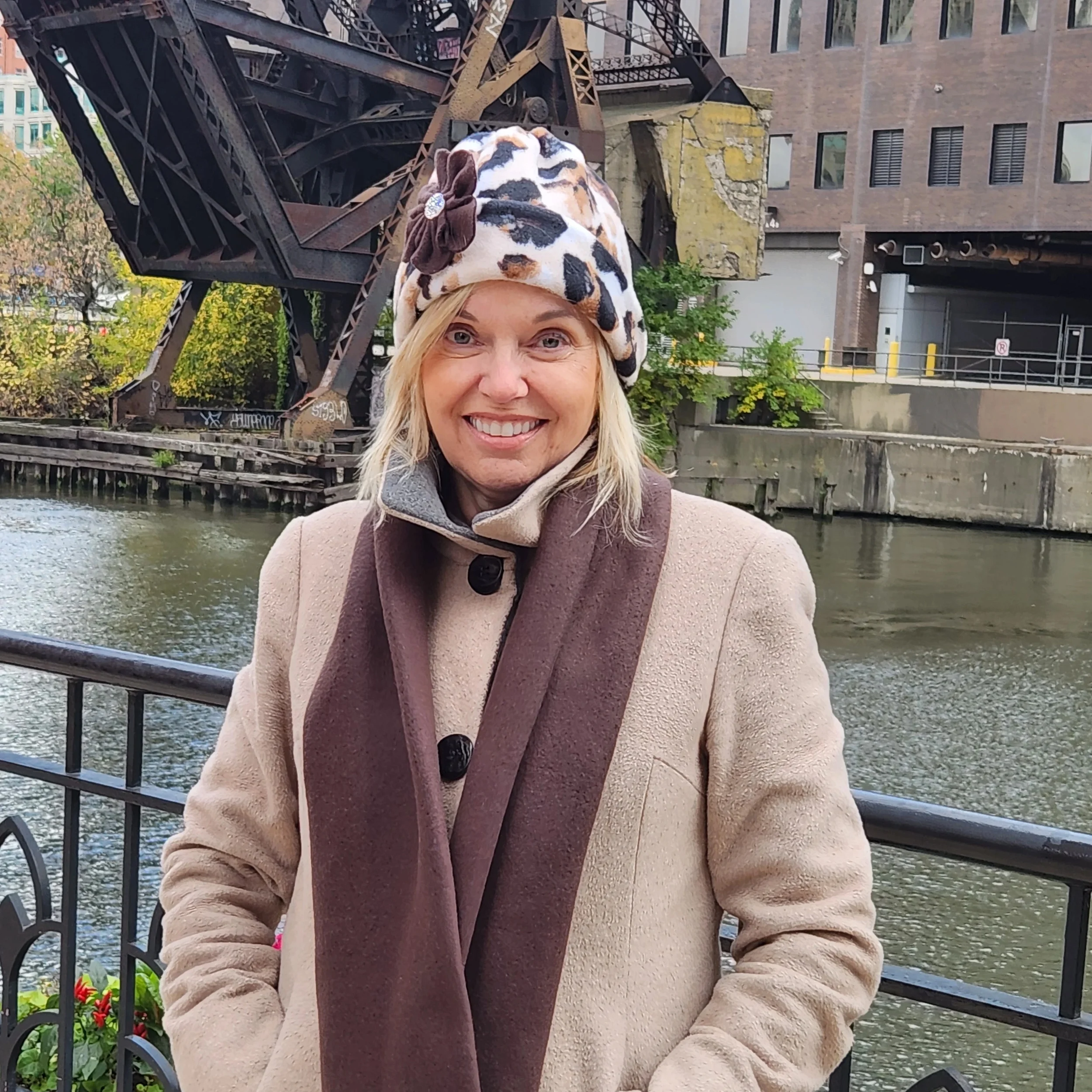 A woman smiling outdoors standing near a waterway, wearing a beige coat, a brown scarf, and a leopard-print hat with a brown flower.