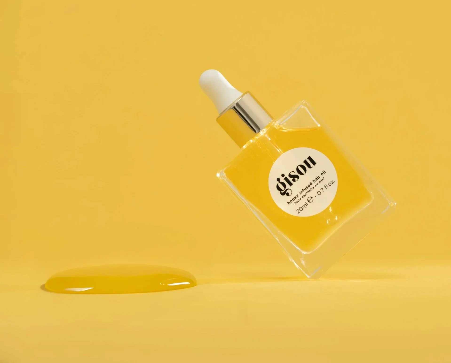A clear glass bottle labeled 'gsoii honey infused hair oil' with a dropper cap, placed diagonally on a yellow background. A small puddle of yellow oil is nearby.