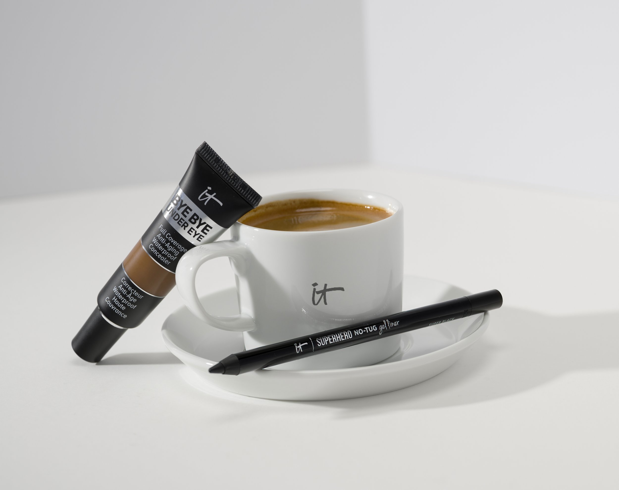 A white mug filled with coffee, a black gel eyeliner pen, and a tube of It Cosmetics Bye Bye Under Eye concealer placed on a white plate on a white surface, with a white background.