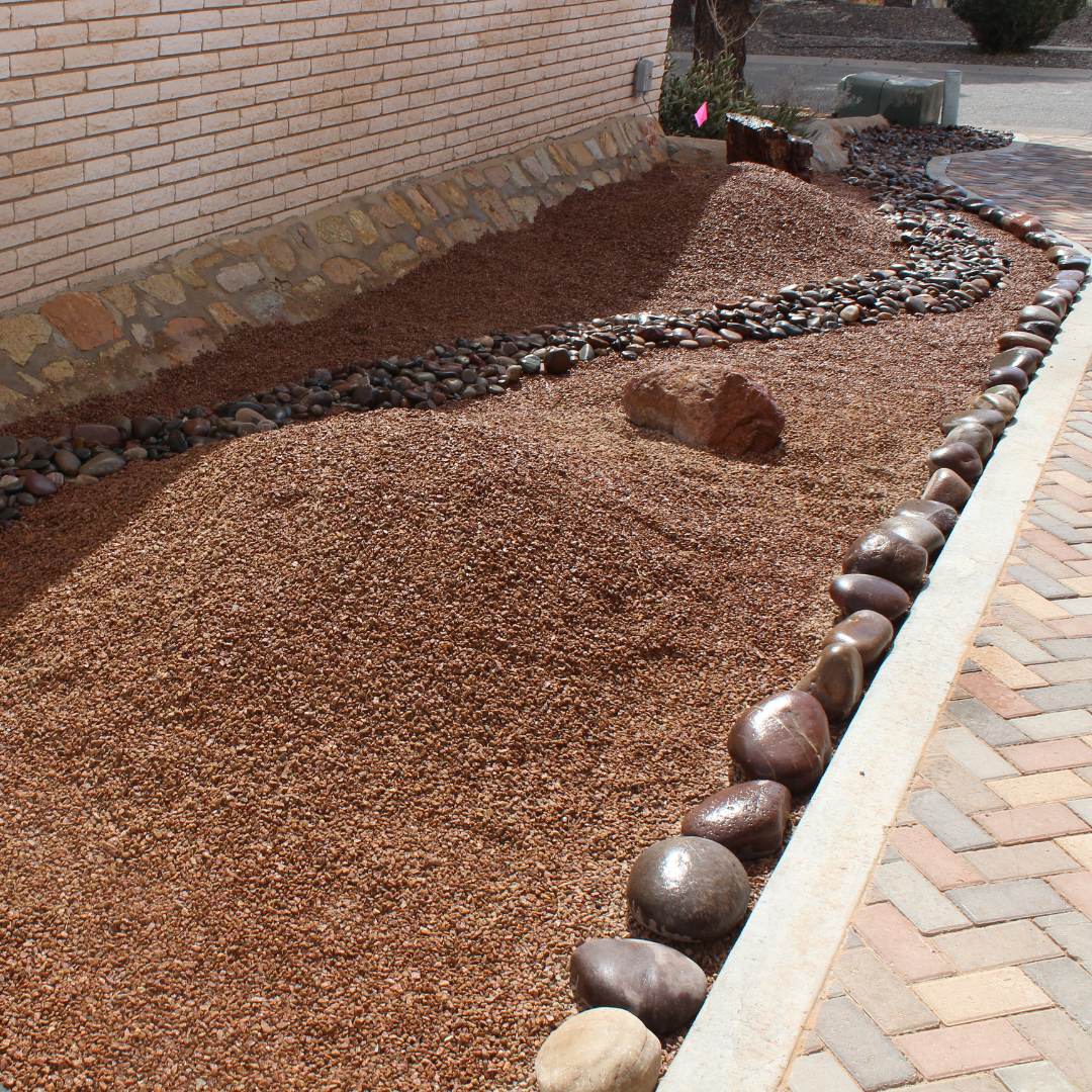 A landscaped area next to a brick building with multicolored rocks creating a border and stacked gravel mounds, surrounded by a brick sidewalk.