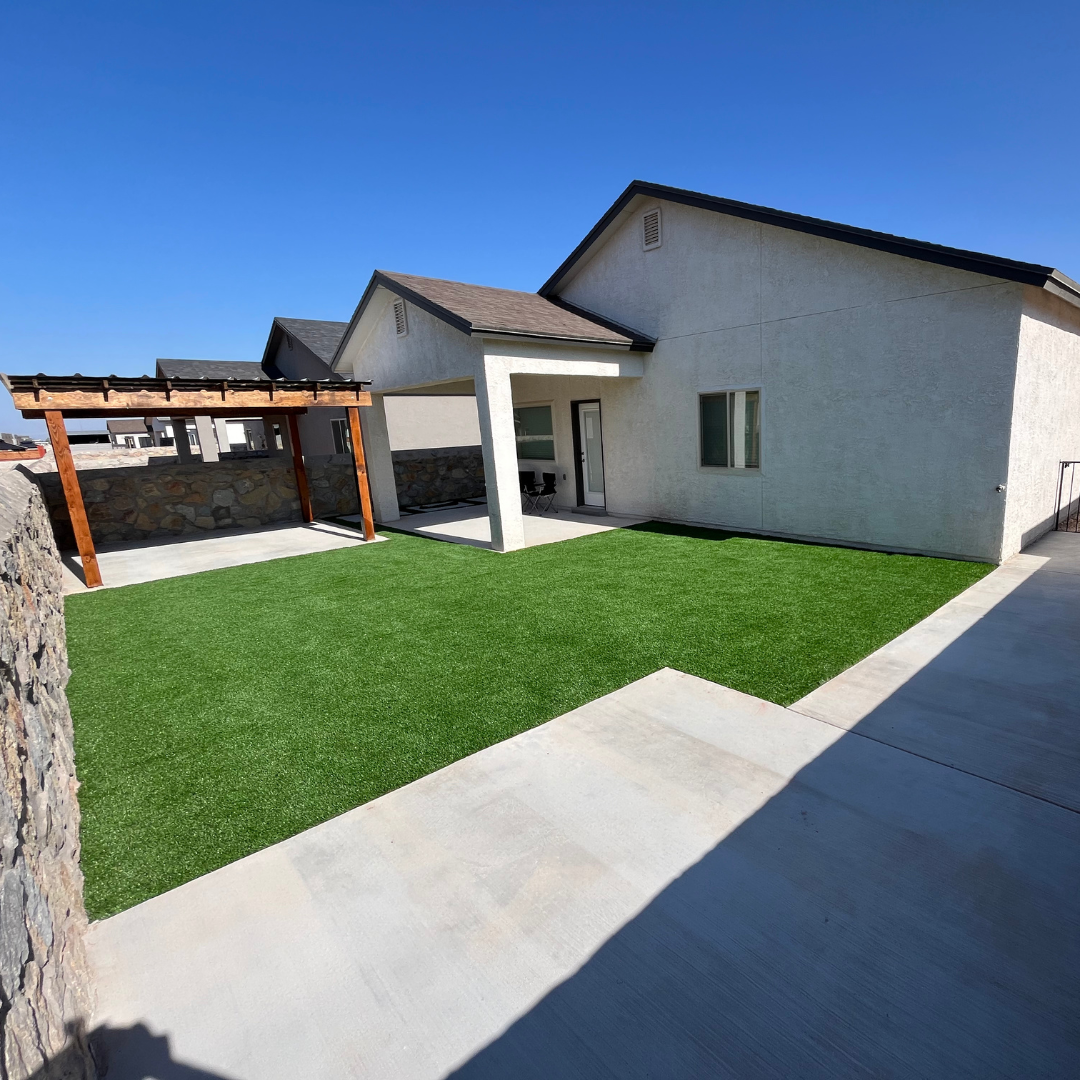 Backyard with green artificial grass, patio with chairs, and modern house with white exterior walls under clear blue sky creating beautiful landscaping.
