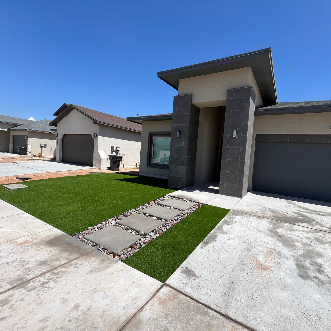 Modern house with a front yard featuring artificial grass, stepping stones, and a driveway with a concrete sidewalk under a clear blue sky.