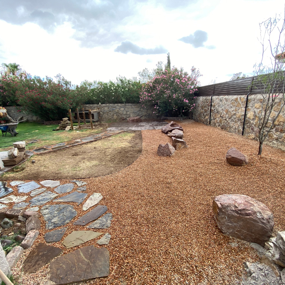 A backyard scene showing a newly landscaped gravel yard with scattered large rocks, a stone pathway, a stone wall, and flowering bushes.