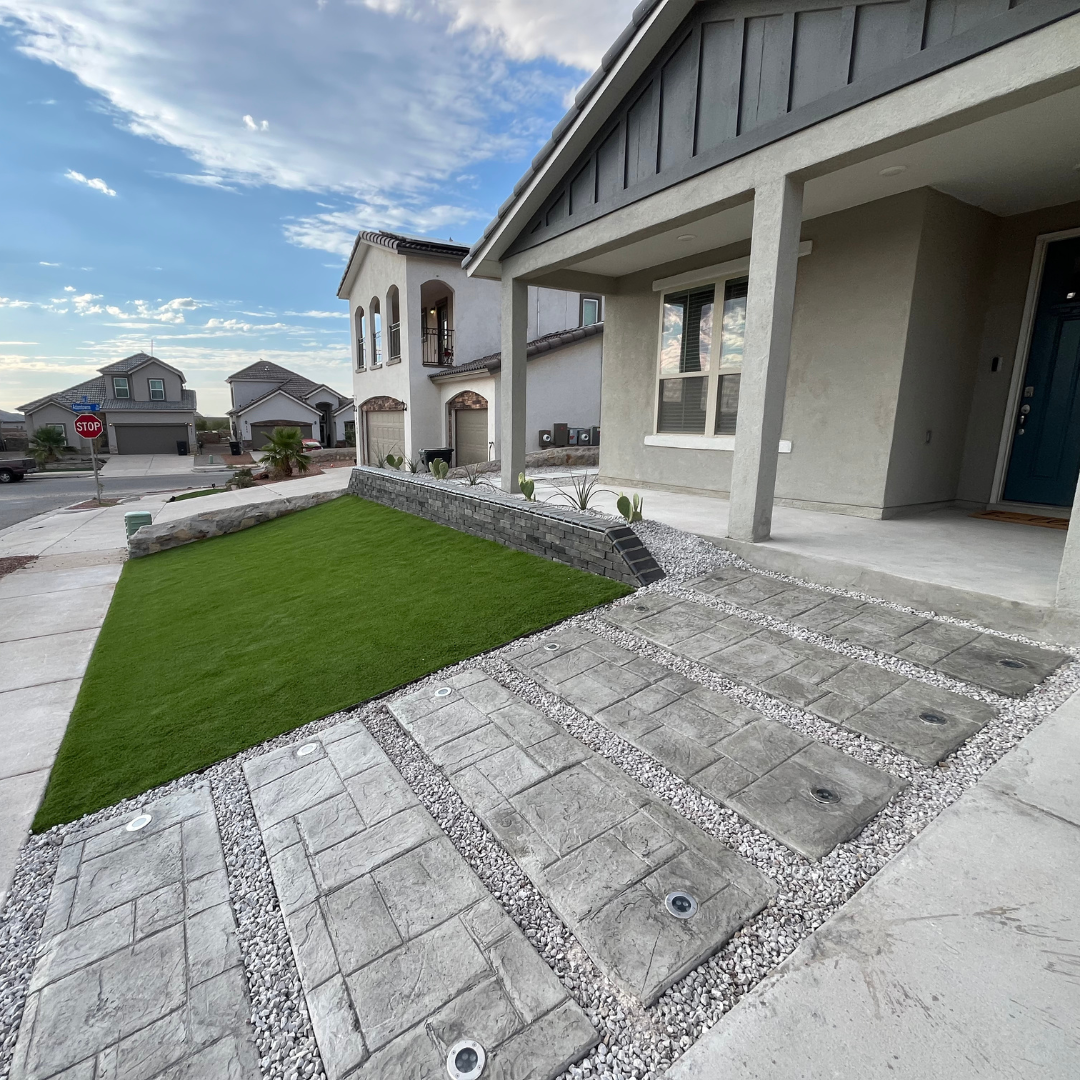 Modern front yard with artificial grass, stone walkway, small cactus plants, and a porch with columns in a suburban neighborhood.