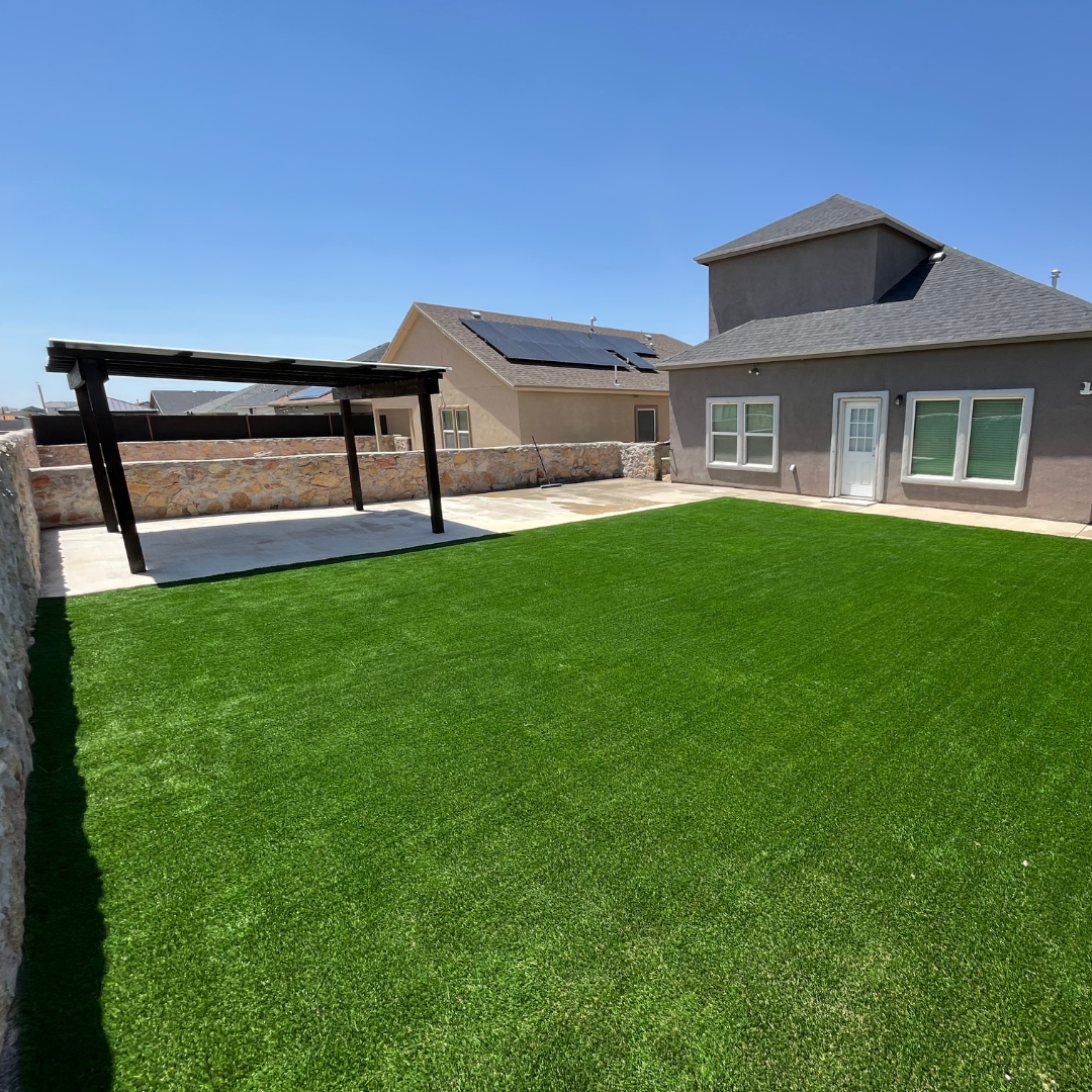 A backyard with a green artificial lawn, a small stone wall, a covered patio, and a house with solar panels on the roof under a clear blue sky.