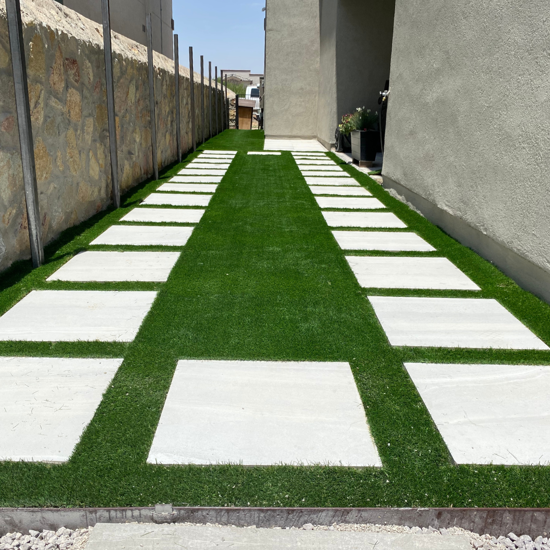 Modern pathway with white rectangular stepping stones and green artificial grass in between, leading along a building with a stone wall on the left and a gray wall on the right.