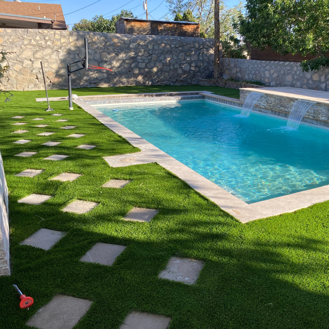 A backyard with a swimming pool with water features, a grass area with stepping stones, a basketball hoop, and a stone wall in the background.