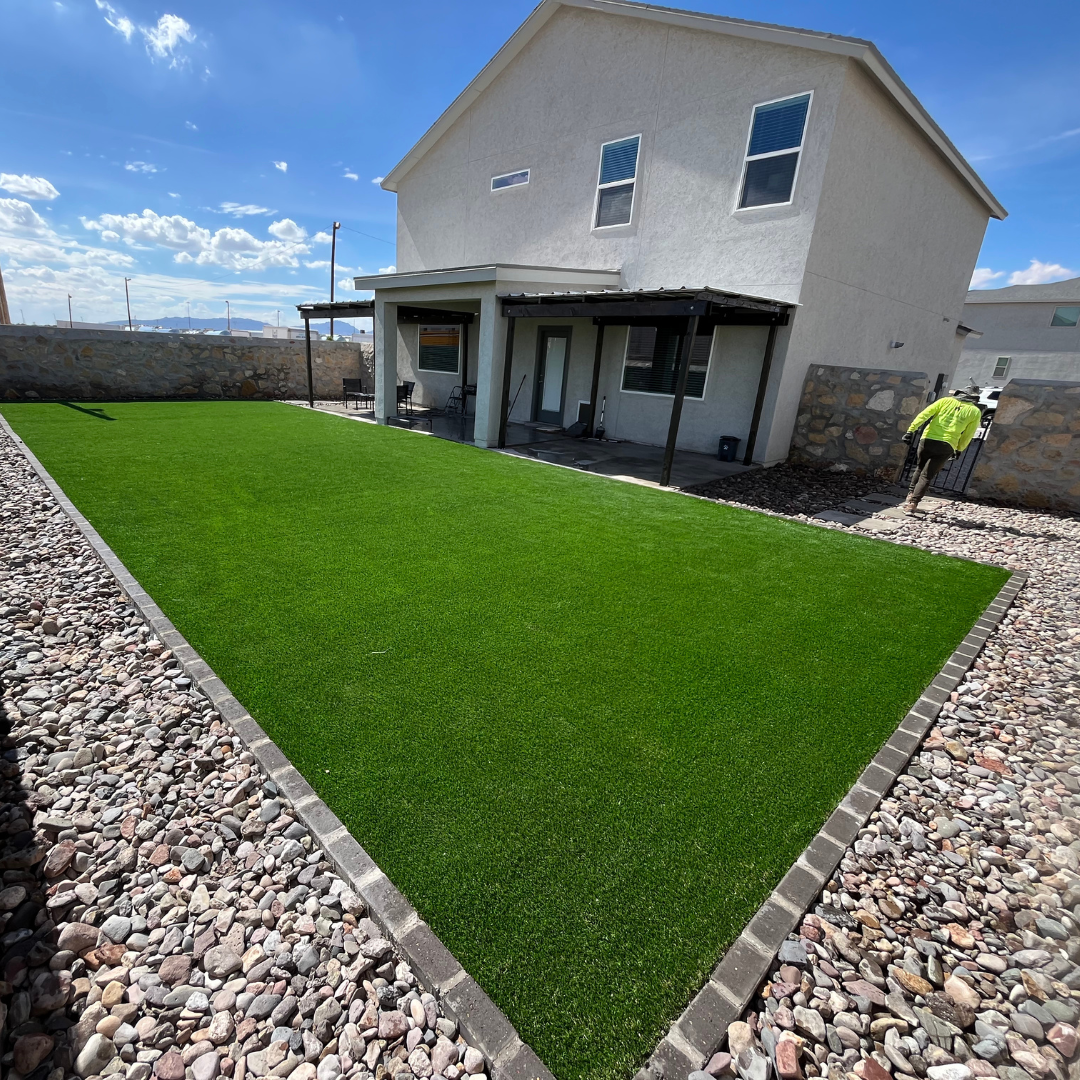 Backyard with a well-maintained green artificial lawn, surrounded by decorative rocks and a stone border, with a two-story house in the background and a person walking near the gate.