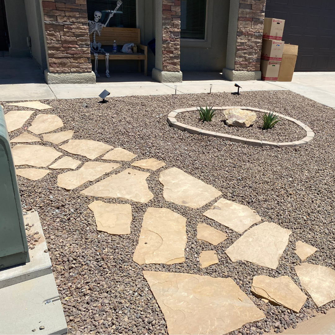 A front yard with a stone pathway leading to the house, a small circular flower bed with two green plants and a large rock, a skeleton figure sitting on a bench under the porch, and cardboard boxes stacked near the garage.
