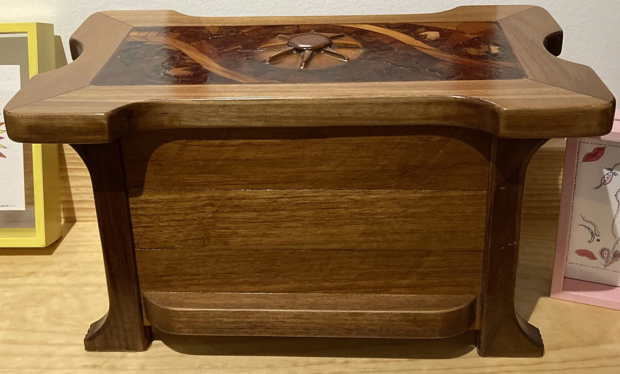 A wooden decorative box with intricate marquetry on the top, sitting on a wooden surface, with partially visible colorful framed artwork on either side.