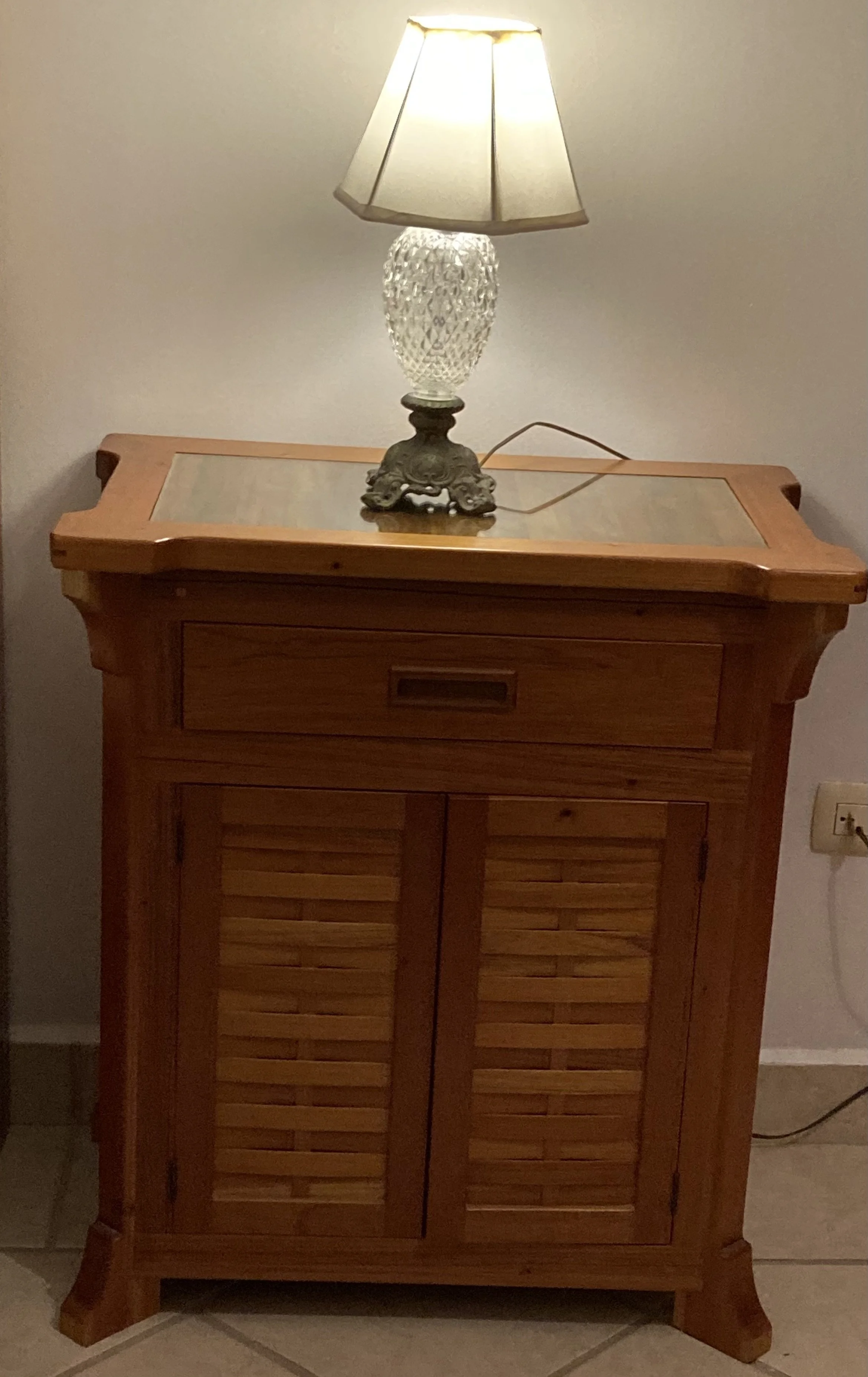 A wooden cabinet with louvered doors and a small drawer on top, with a decorative bedside lamp on it. The lamp has a crystal base and a beige lampshade, plugged into an electrical outlet.