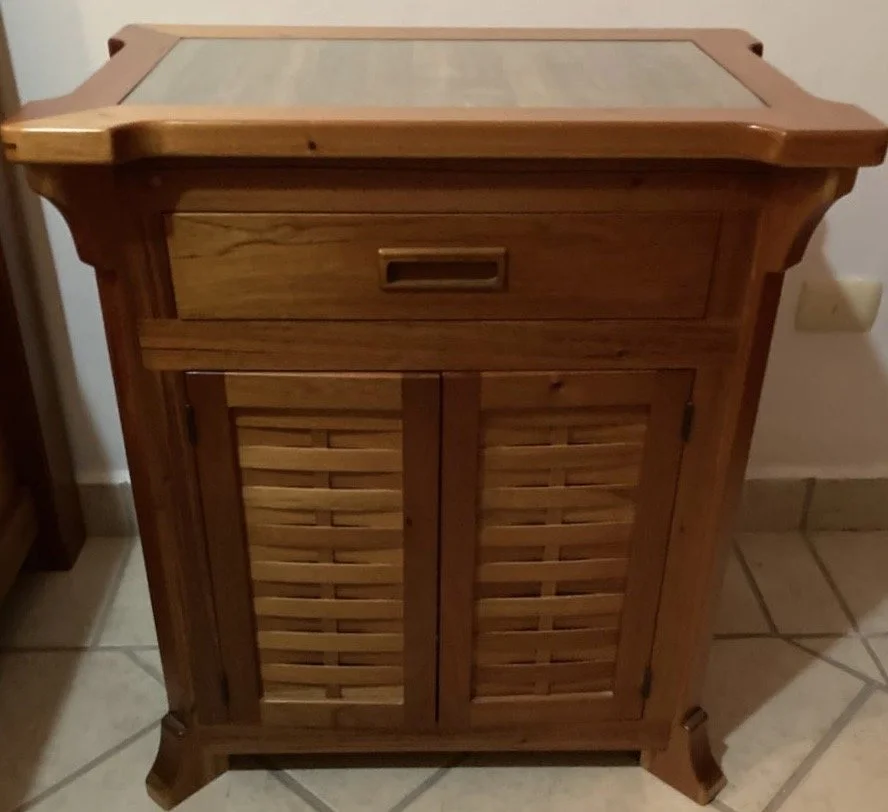 A wooden cabinet with a drawer on top and two louvered doors below, standing on a tiled floor.