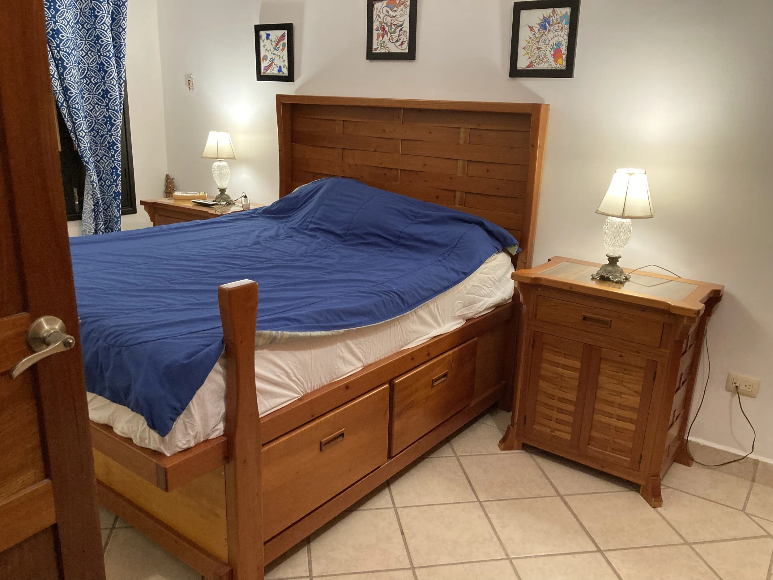 A bedroom with a wooden bed and headboard, a blue bedspread, and two wooden nightstands with lamps, decorated with framed artwork on the wall.
