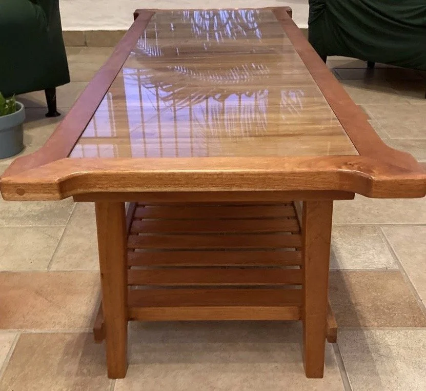A wooden coffee table with a glass top and slatted shelf underneath, located in a room with tiled flooring and plants.