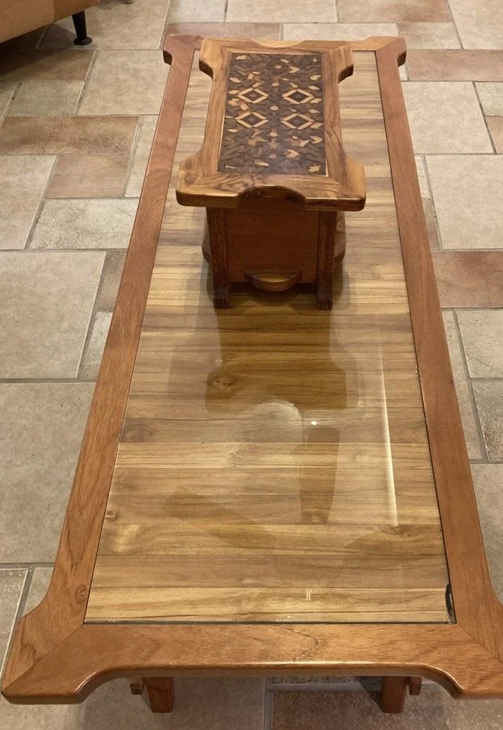A wooden table with a glass top and a smaller wooden table with intricate inlay design, placed on a tiled floor.