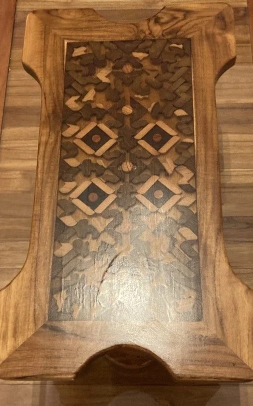 Top view of a wooden sandbox with intricate geometric inlay design in the center, surrounded by a thick wooden border, on a wooden floor.