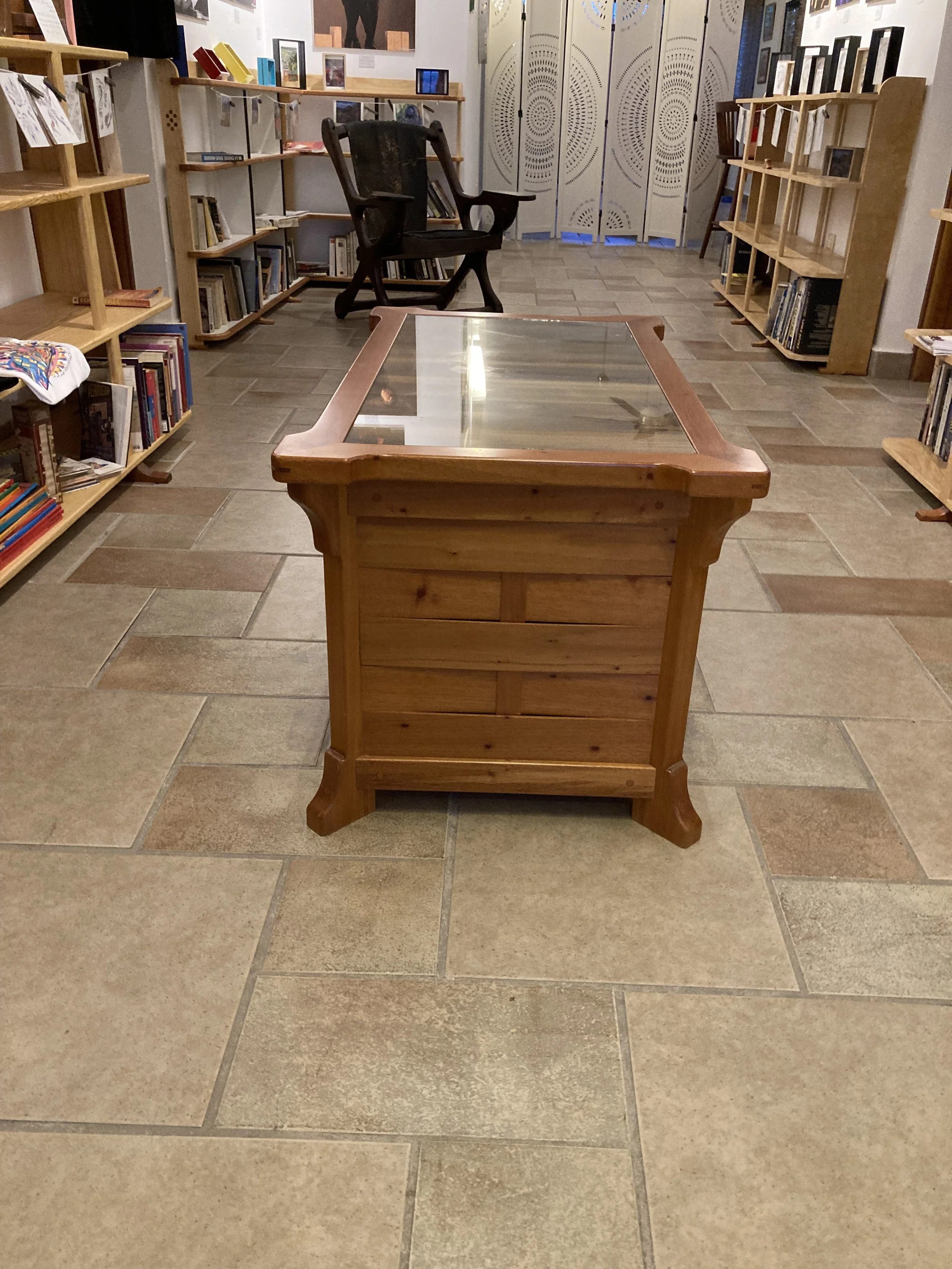 A wooden display table with a glass top in the center of a bookstore or library. Behind it is a black armchair surrounded by bookshelves filled with books and framed pictures on top. The room has tile flooring and decorative folding doors in the back