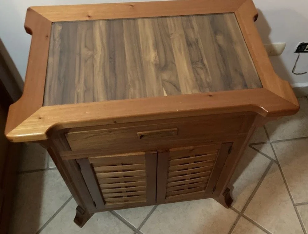 A wooden side table with a glass top, slatted cabinet door, sitting on a tiled floor next to a wall and an electrical outlet.