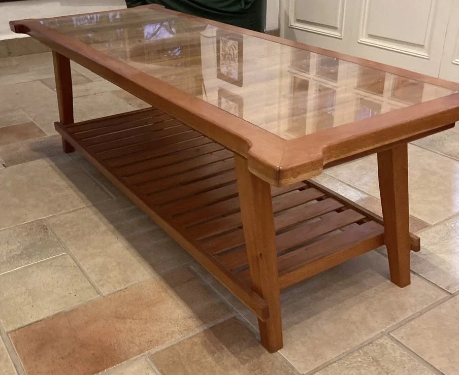 A wooden coffee table with a glass top and a lower slatted shelf, placed on a tiled floor in a room with white cabinetry.