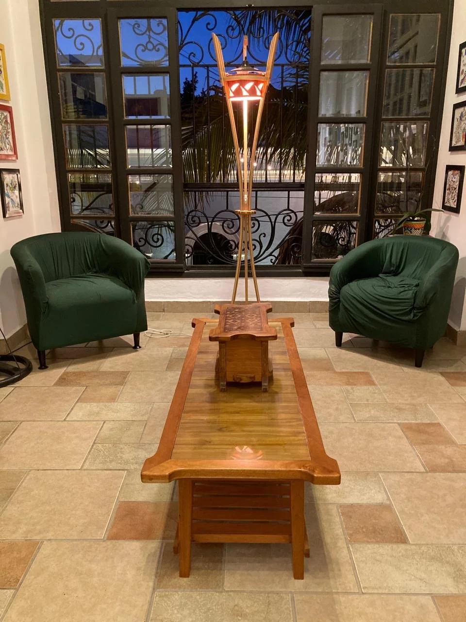 A cozy living room scene featuring a unique floor lamp with a face design, two green armchairs, a wooden coffee table, and a large window with decorative ironwork, viewing a darkening sky outside.