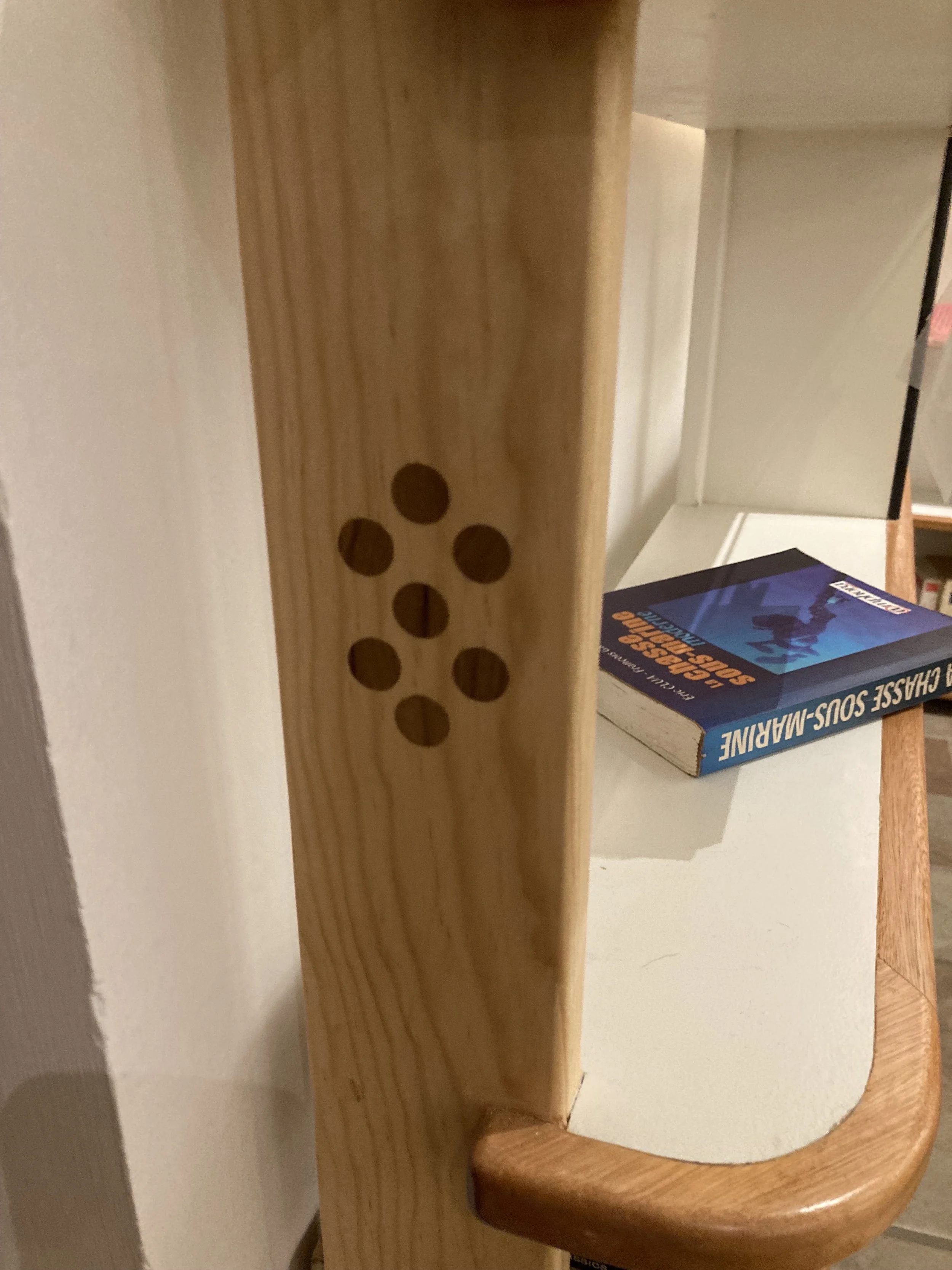 Close-up of a piece of furniture with a wooden vertical support with several round holes, next to a white surface, with a book titled 'The Chase Sou-Marine' resting on top.