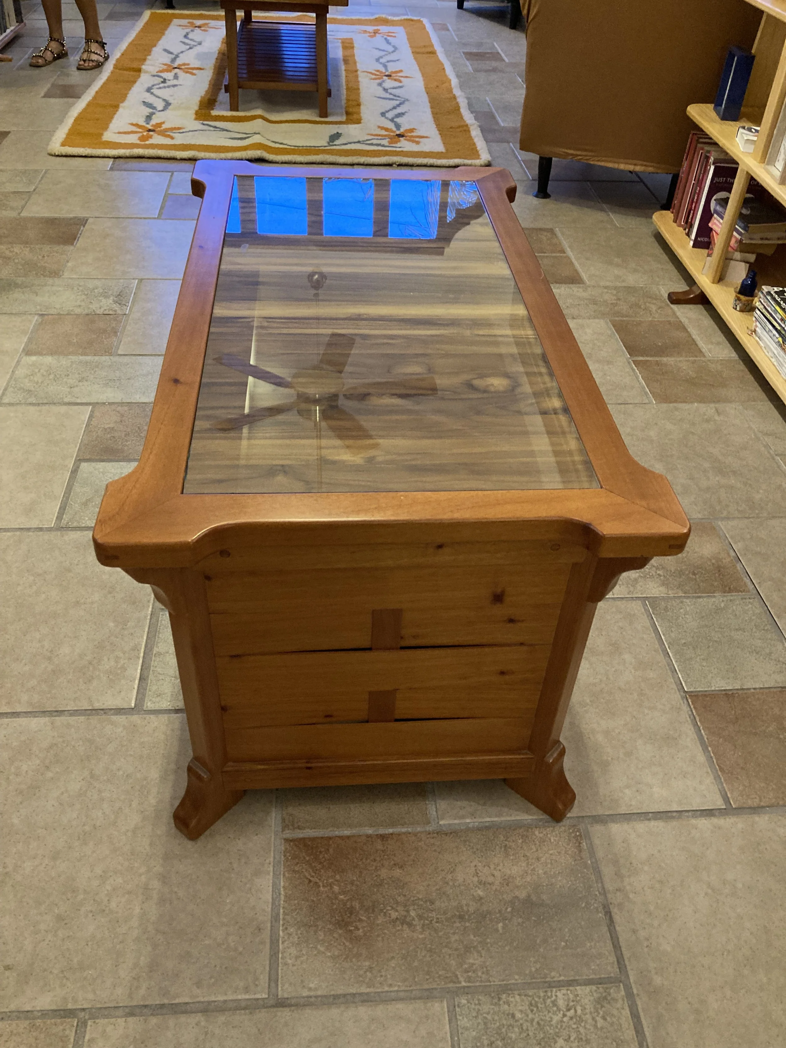 A wooden coffee table with a glass top, situated on a tiled floor inside a home. The glass reveals the wooden pattern underneath. In the background, there is a patterned area rug with a small table on it and part of a person’s legs and feet are visib