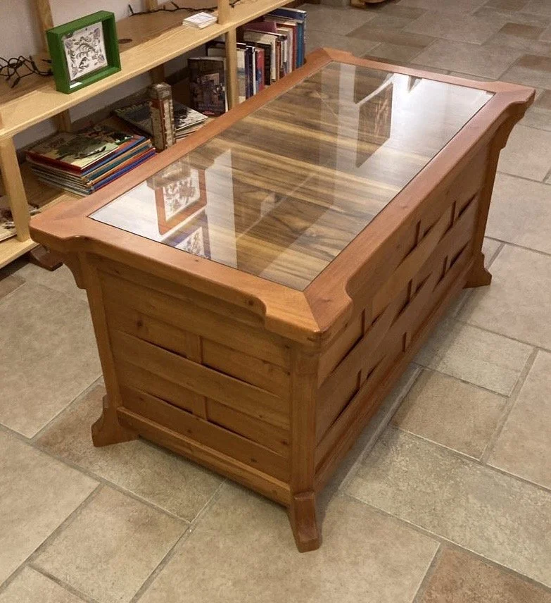 Wooden coffee table with a glass top in a room with tile flooring and a bookshelf in the background.