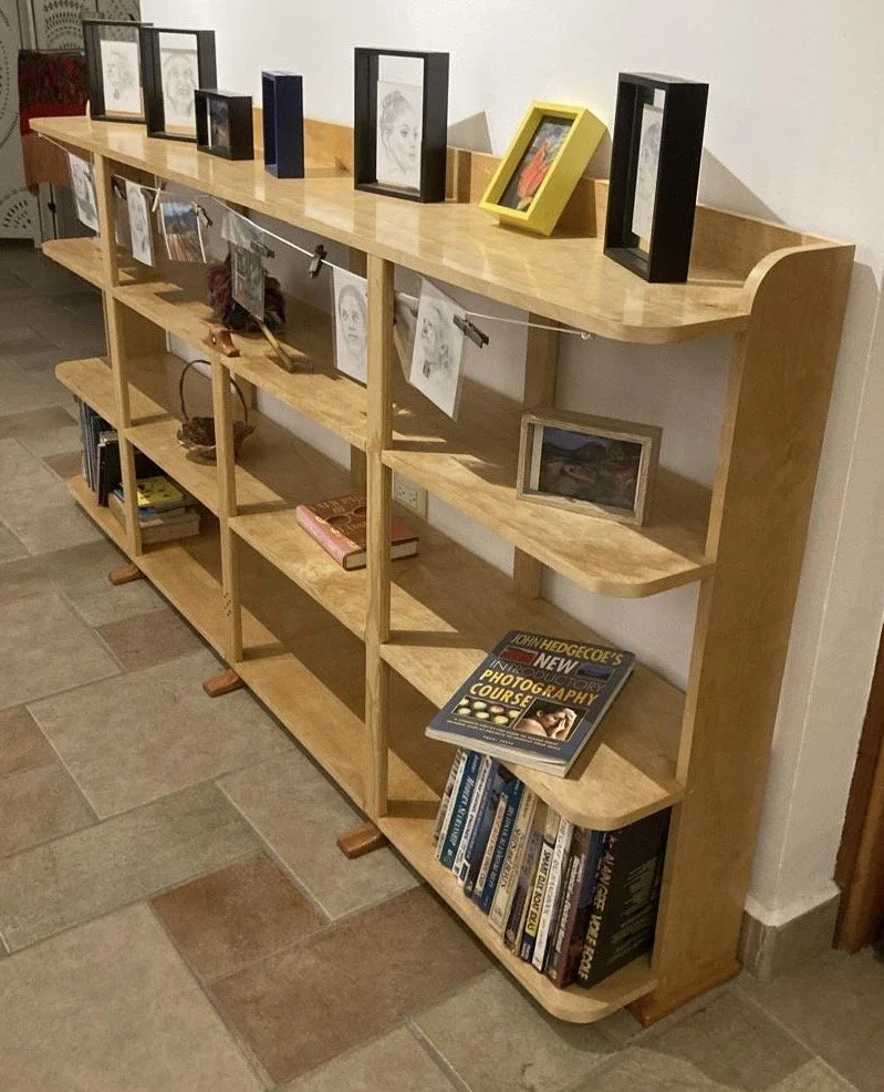 A wooden bookshelf with three shelves filled with books, framed pictures, and artwork, with additional smaller frames and a book on photography on top.