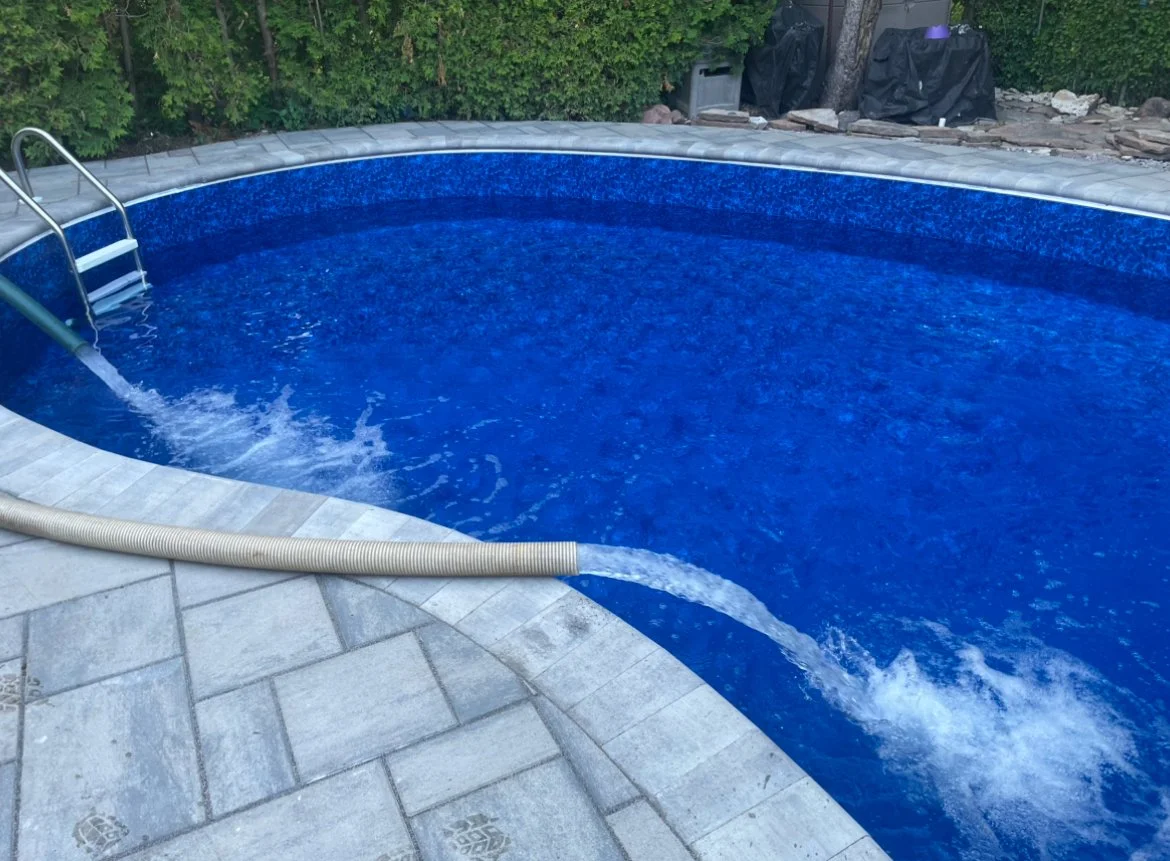 Inground swimming pool with blue interior, surrounded by a stone patio, with water flowing in from a hose, and dense bushes in the background.