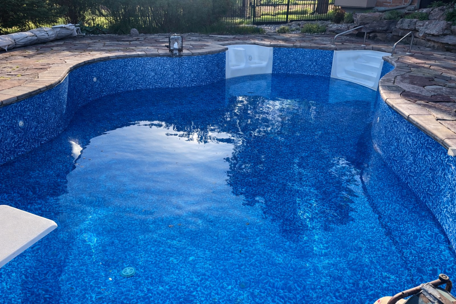 An empty swimming pool with a blue mosaic interior, surrounded by a stone deck, with a metallic handrail on one side and a white seat in the foreground.