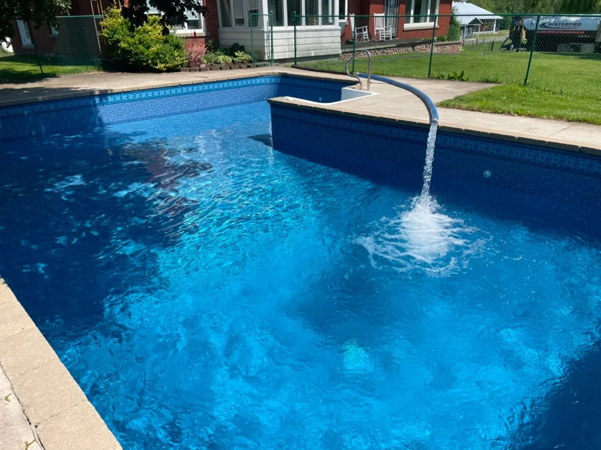 Empty in-ground swimming pool with water feature pouring water into it, surrounded by concrete deck and a grassy yard, with a chain-link fence and house in the background.