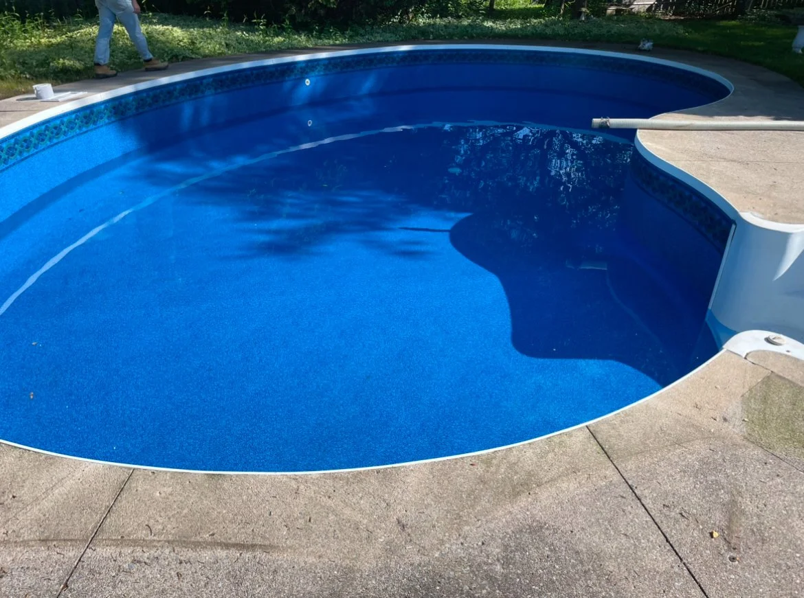 Empty blue in-ground swimming pool with a lifting pole resting along the edge, surrounded by a concrete deck and some greenery in the background.