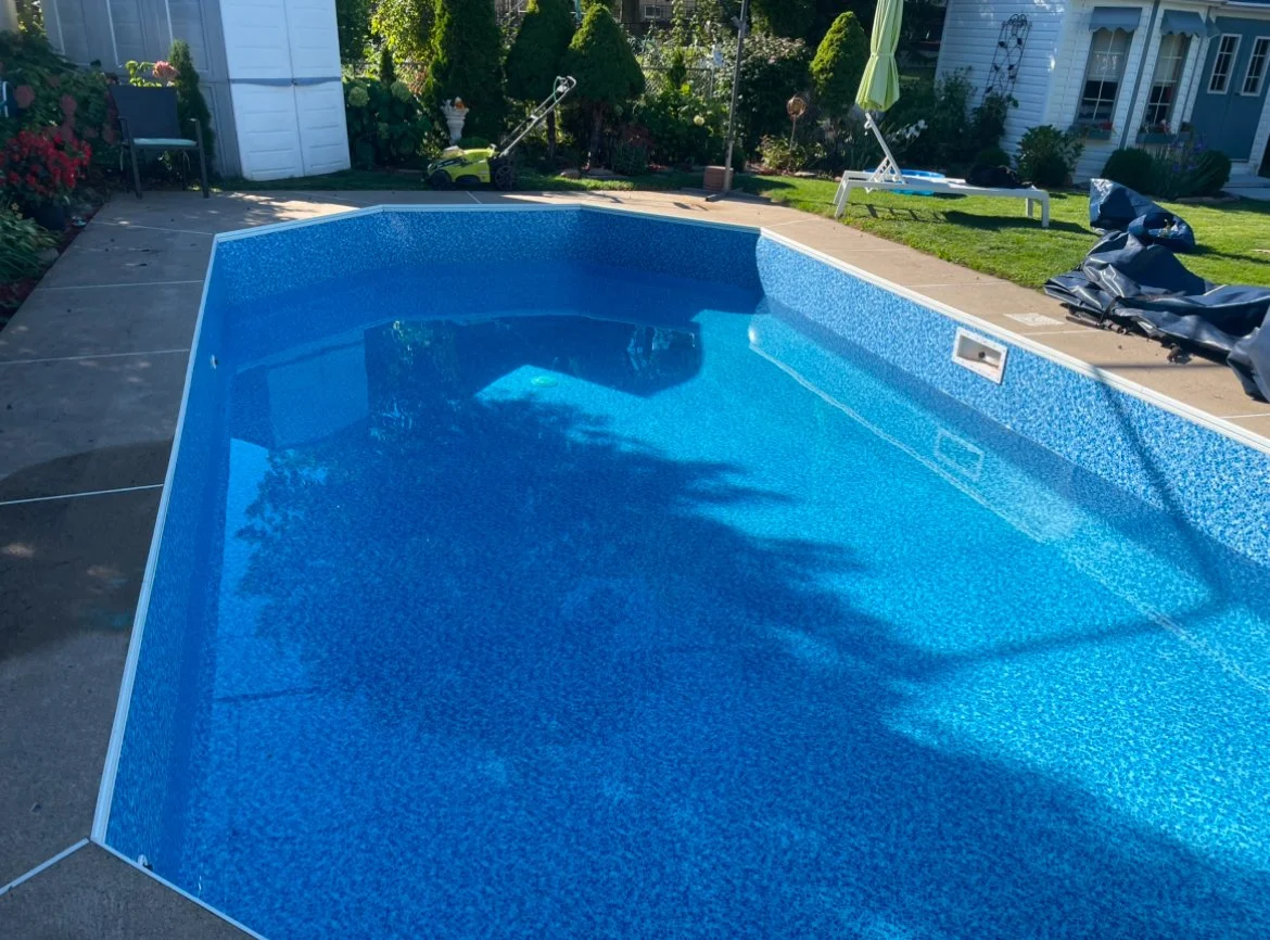 Empty in-ground swimming pool with blue mosaic tiles, surrounded by a concrete deck and garden area with outdoor furniture and umbrellas on a sunny day.
