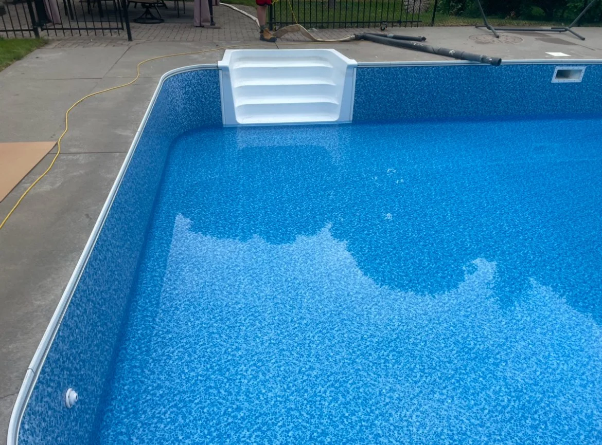 Close-up view of a newly installed in-ground swimming pool with blue speckled interior and white steps at the shallow end, surrounded by concrete deck.