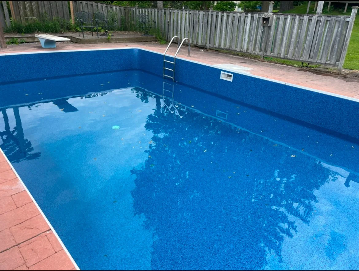 Empty backyard swimming pool with metal ladder, surrounded by brick patio, wooden fence, and some outdoor furniture.