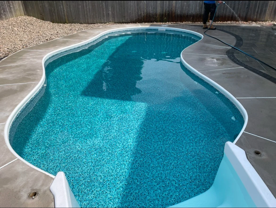 In-ground swimming pool with a curved shape, filled with blue water, surrounded by a concrete deck, with a person cleaning with a hose in the background.