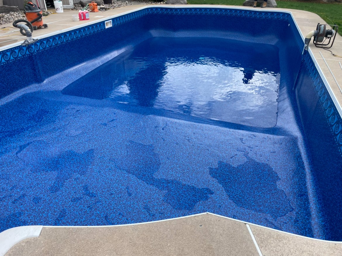 An empty in-ground swimming pool with blue lining and a small amount of water at the bottom, located on a concrete deck with some cleaning and maintenance equipment around it.