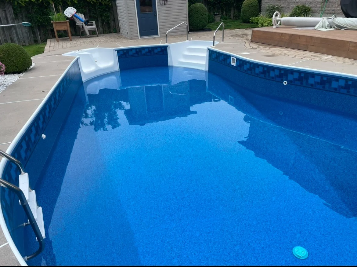 Empty in-ground swimming pool with blue lining, metal handrails at the steps, surrounded by a concrete deck, with backyard landscaping including bushes, a shed, and outdoor furniture.