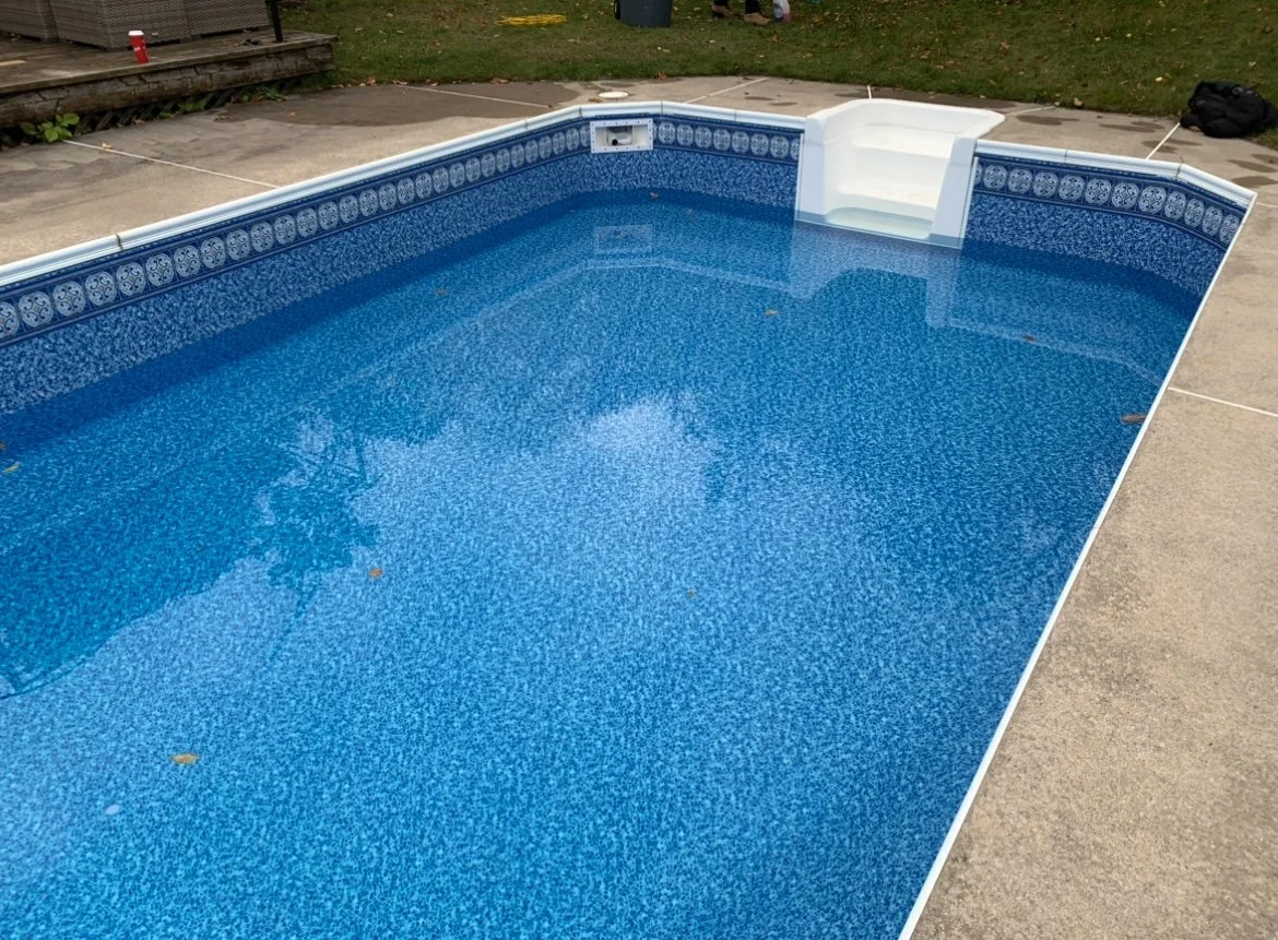 Empty swimming pool with blue tile interior and a small white stair at the far end, surrounded by a concrete deck and a grassy backyard.