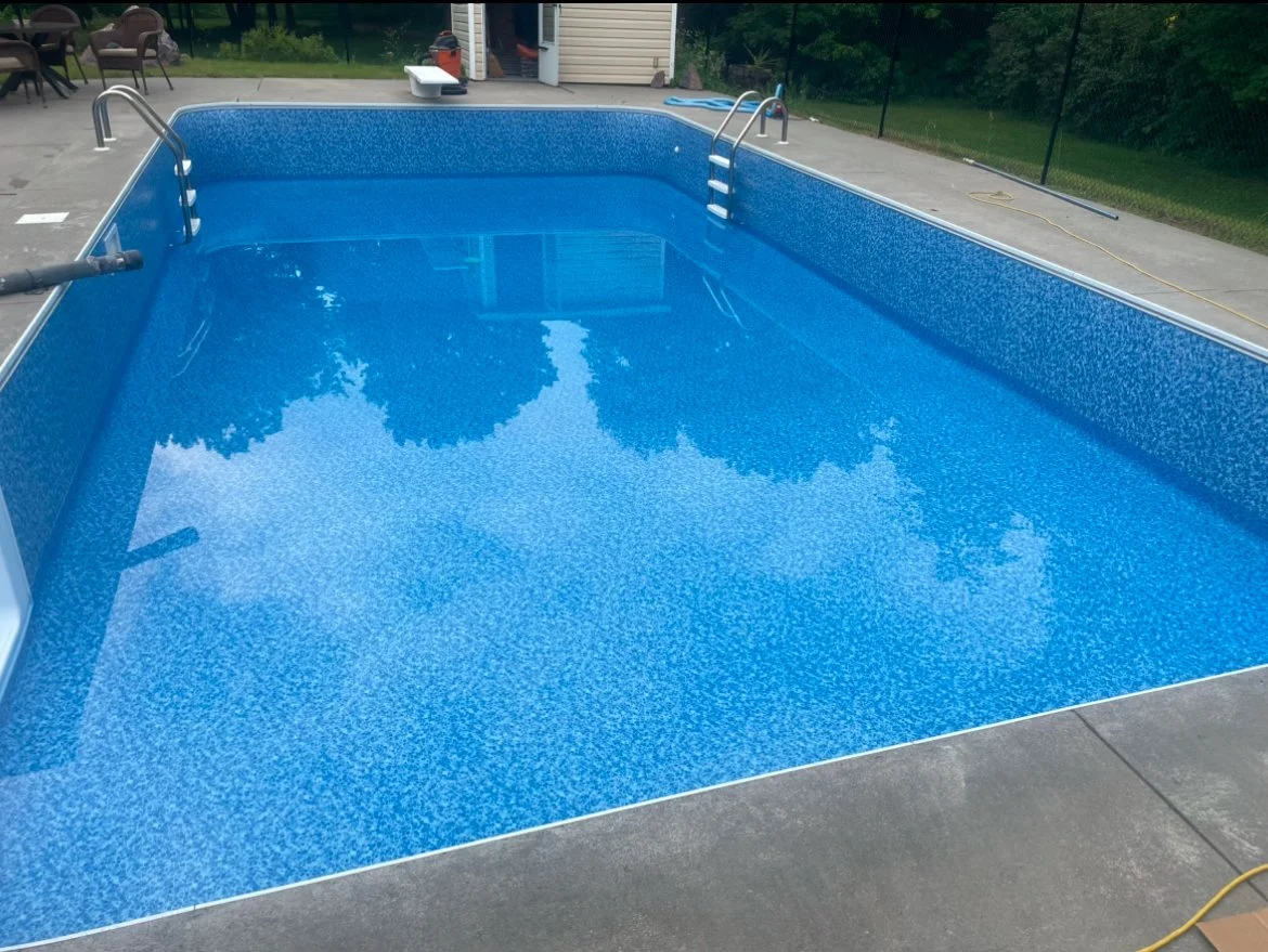 Empty in-ground swimming pool with blue tile interior, metal ladders, and a concrete deck, surrounded by outdoor furniture and a grassy backyard.