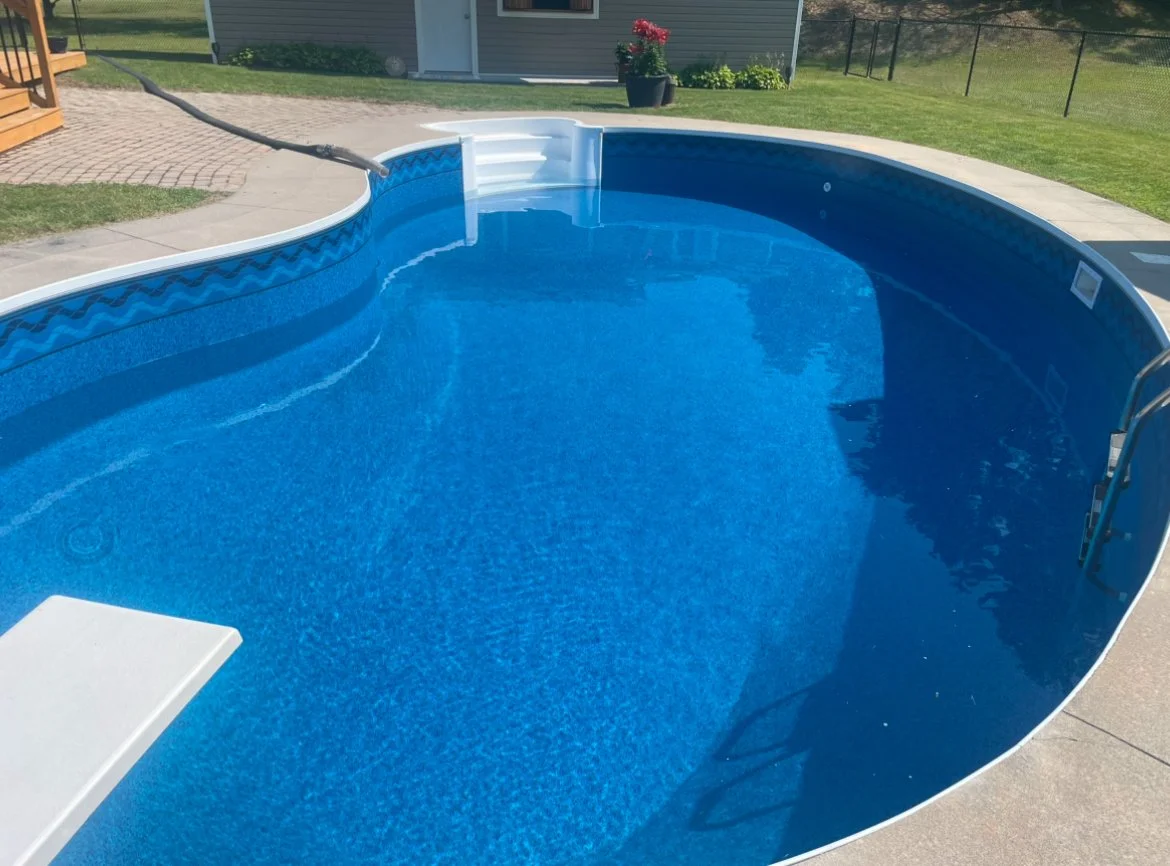 Above-ground swimming pool filled with blue water in a backyard with concrete and grass, a house in the background, and potted flowers.