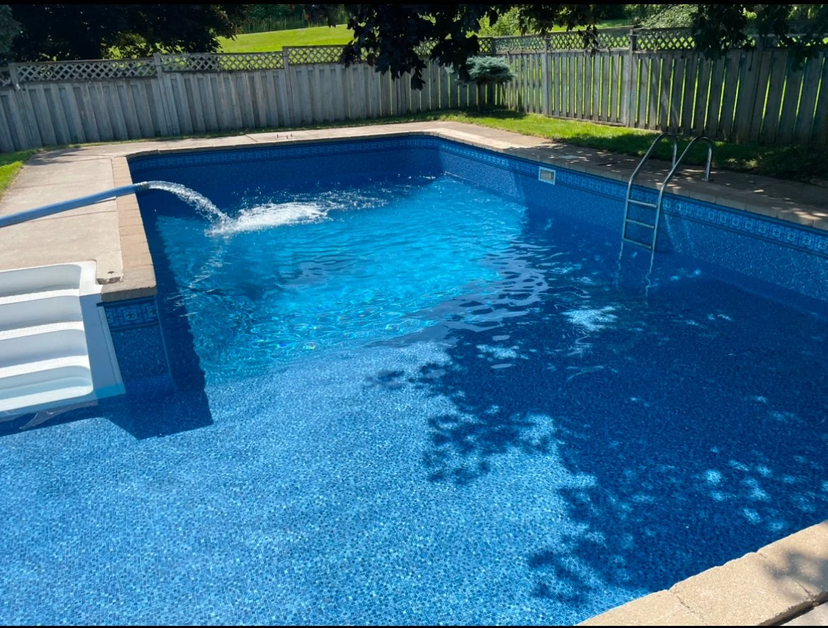 Underground swimming pool with blue tiles, a ladder, and a pool cover at the edge surrounded by a wooden fence and green lawn.