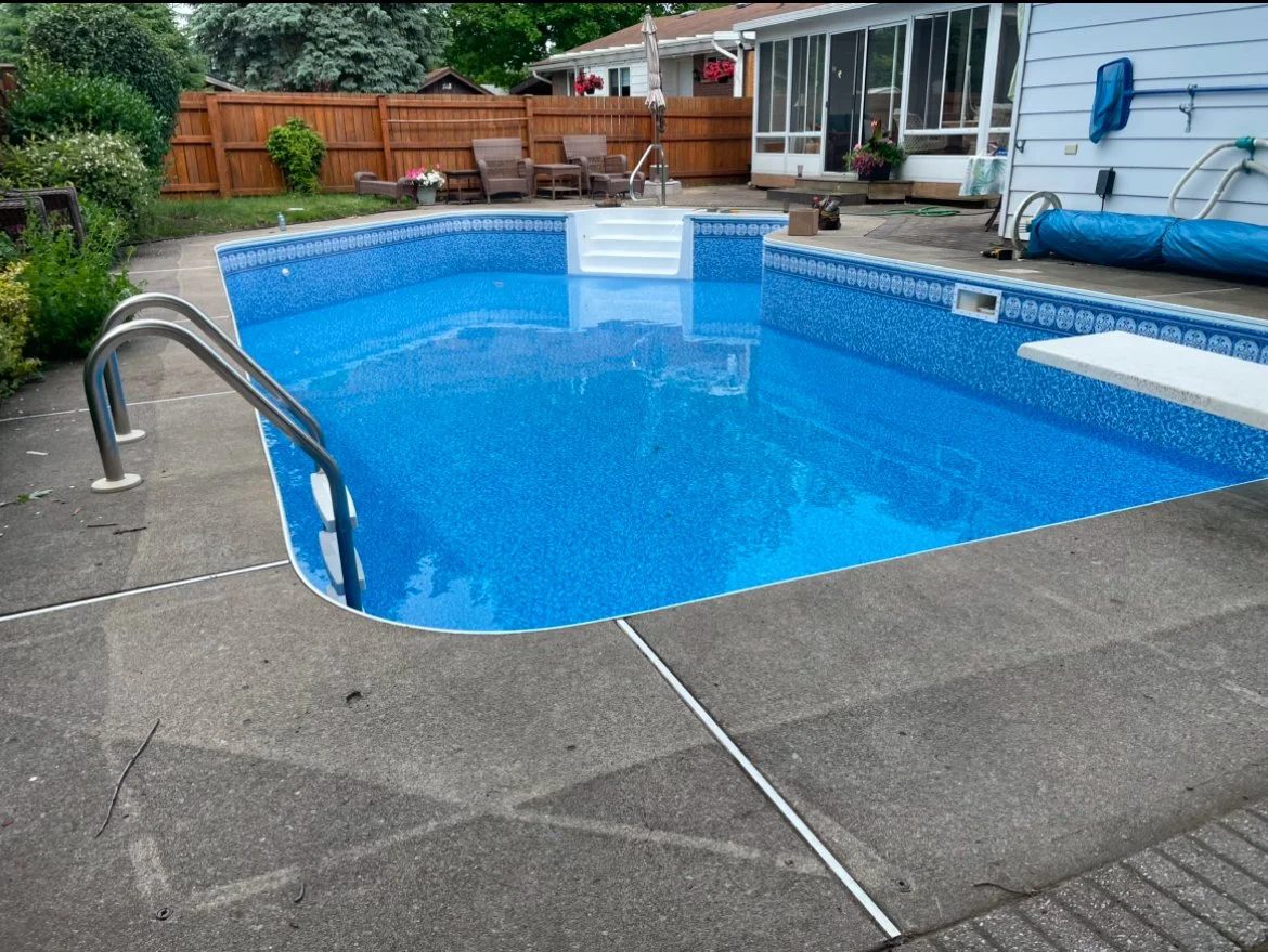 An outdoor backyard swimming pool with a blue interior and steps leading into the water, surrounded by a concrete deck, with a wooden fence and garden furniture in the background.