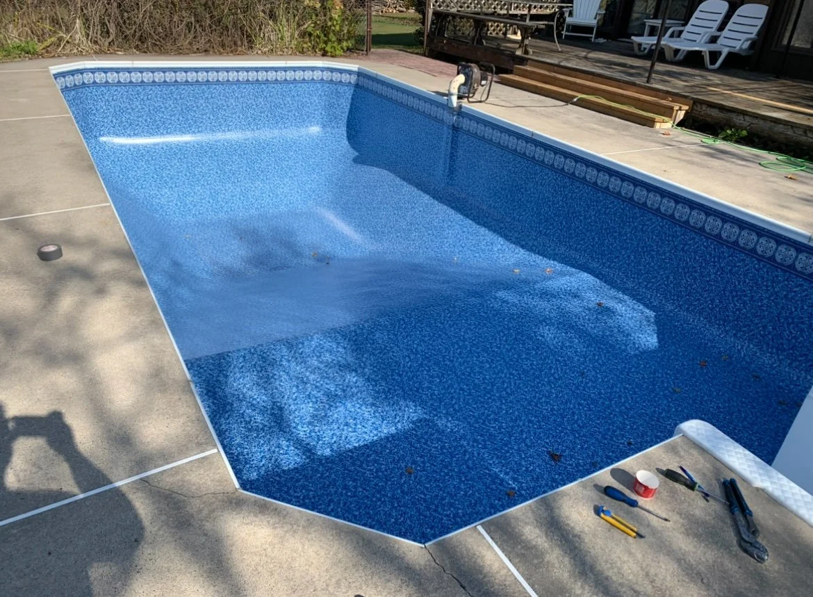 An empty in-ground swimming pool with blue patterned interior tiles, surrounding concrete deck, and tools for pool maintenance beside it. In the background, there is a wooden deck with outdoor chairs.
