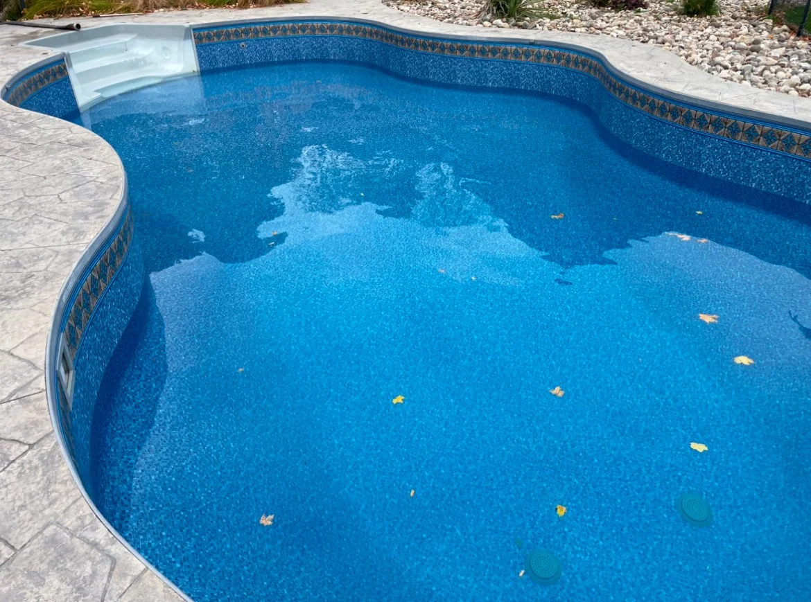 Empty in-ground swimming pool with blue water, surrounded by gray stone patio and decorative tile border, with some fallen leaves floating on the surface.