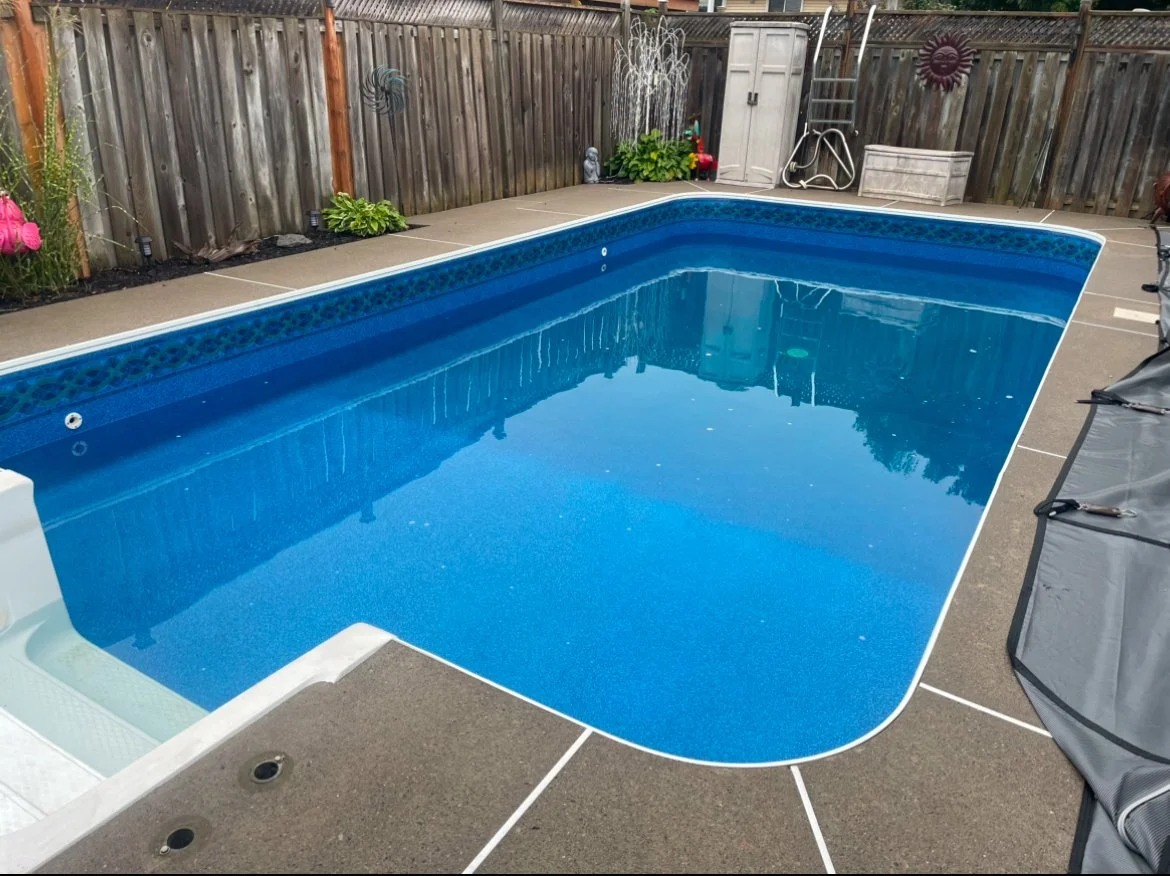 An empty backyard swimming pool with a blue interior, surrounded by a concrete pool deck and wooden fence. Pool cover is pushed to the side, and pool equipment is visible.