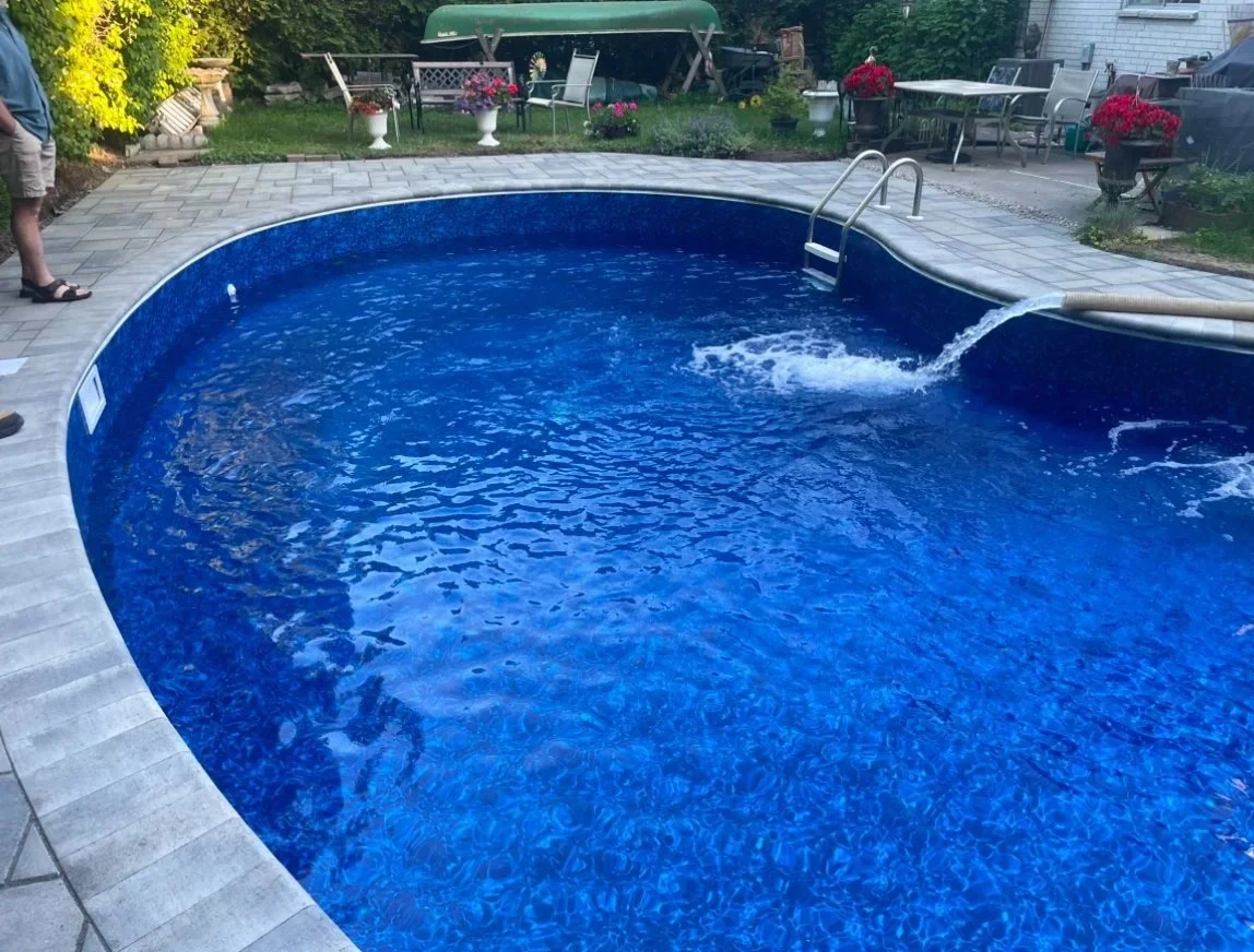 Backyard with an in-ground blue swimming pool, surrounded by a paved area, patio furniture, and potted flowers. A person is partially visible on the left side.